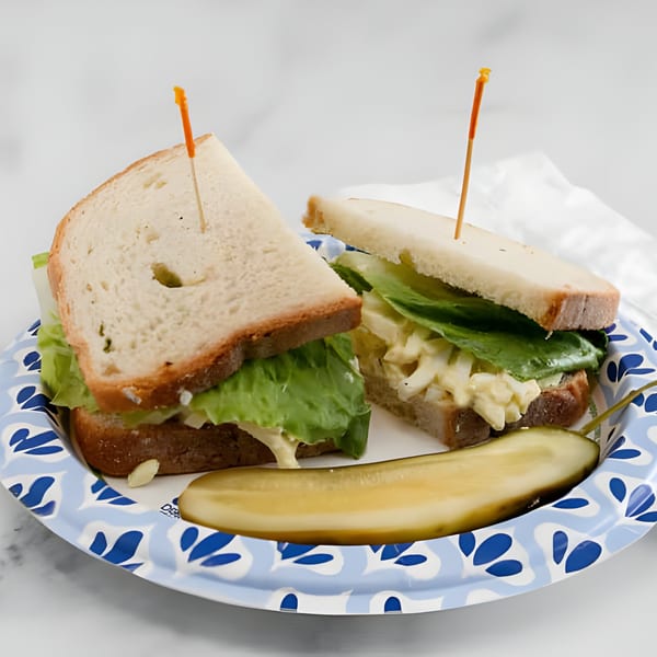 Two egg salad sandwiches with lettuce and a pickle spear on a blue and white paper plate.