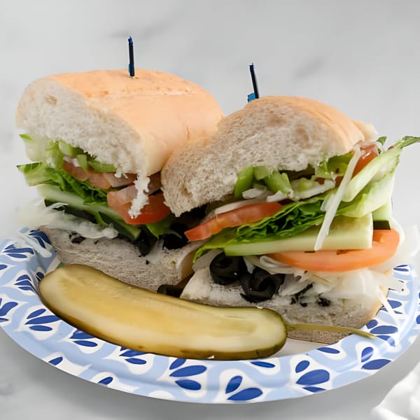 Two Mo's Deli sandwiches with lettuce, tomato, cucumber, olives, and a pickle spear on a plate.