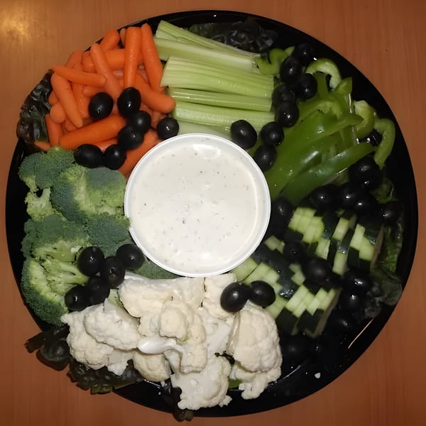 A black platter of fresh vegetables including carrots, celery, broccoli, cauliflower, cucumbers, and olives with a side of ranch dip.