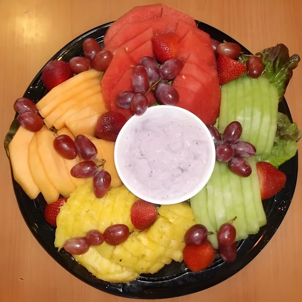 A black platter of fresh fruit, including watermelon, cantaloupe, pineapple, grapes, strawberries, and honeydew, with a small cup of dip.