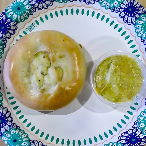 A Mo's Deli bagel with cream cheese and a side of green pesto on a decorative paper plate.